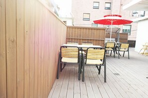 A wooden deck with a table and chairs, a red umbrella, and a building in the background.