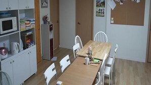 A dining area with a wooden table, white chairs, a white cabinet with kitchen appliances, and a corkboard with photos on the wall. - Agit Guesthouse (Seoul)