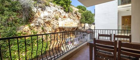 A balcony with a view of a rocky hillside and greenery. There are wooden chairs and a table on the balcony.