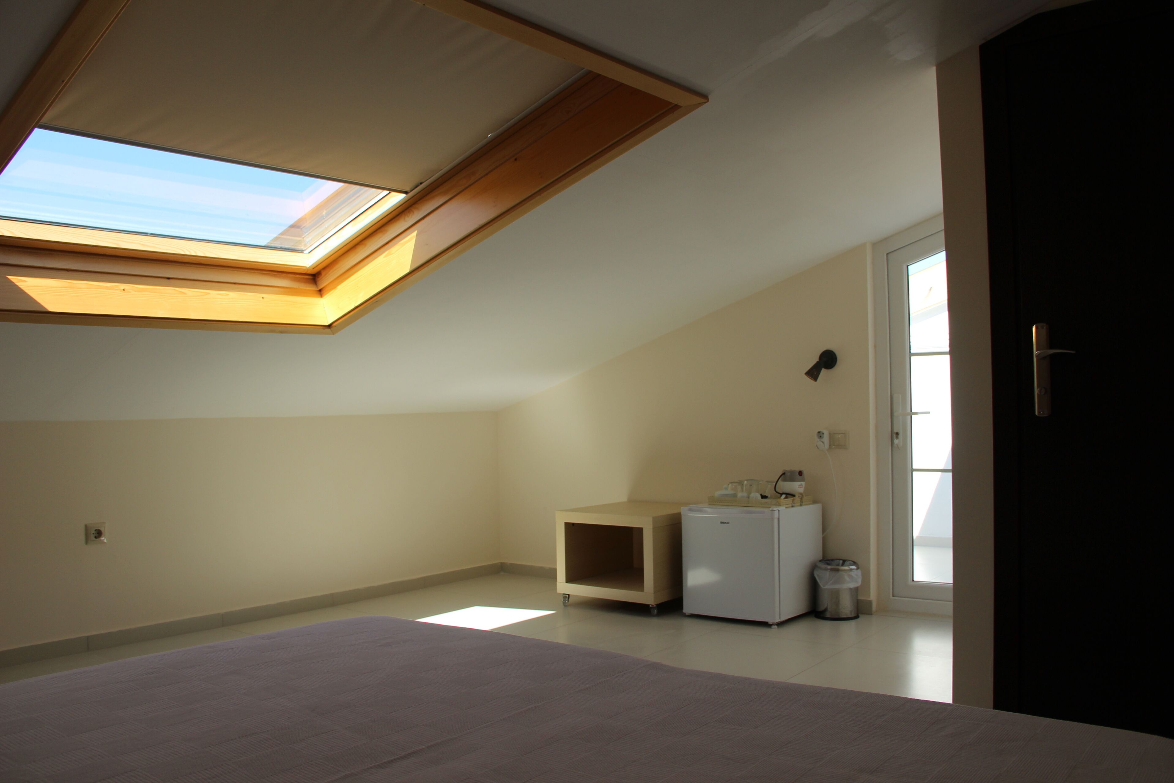 Une chambre avec un puits de lumière, un lit, une petite armoire blanche et une porte donnant sur une autre pièce.
