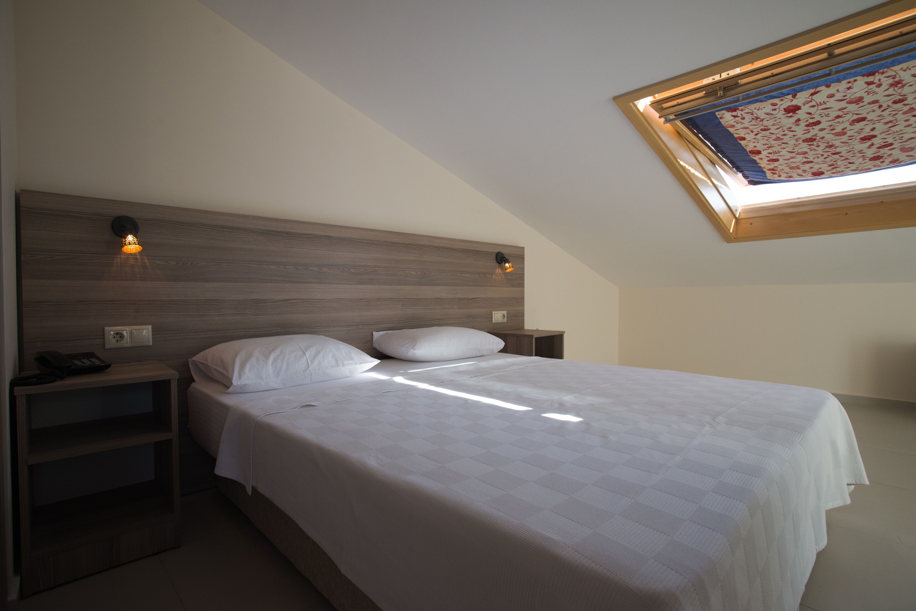 A bedroom with a wooden headboard, a single bed with white bedding, two bedside tables, and a skylight with a floral view.