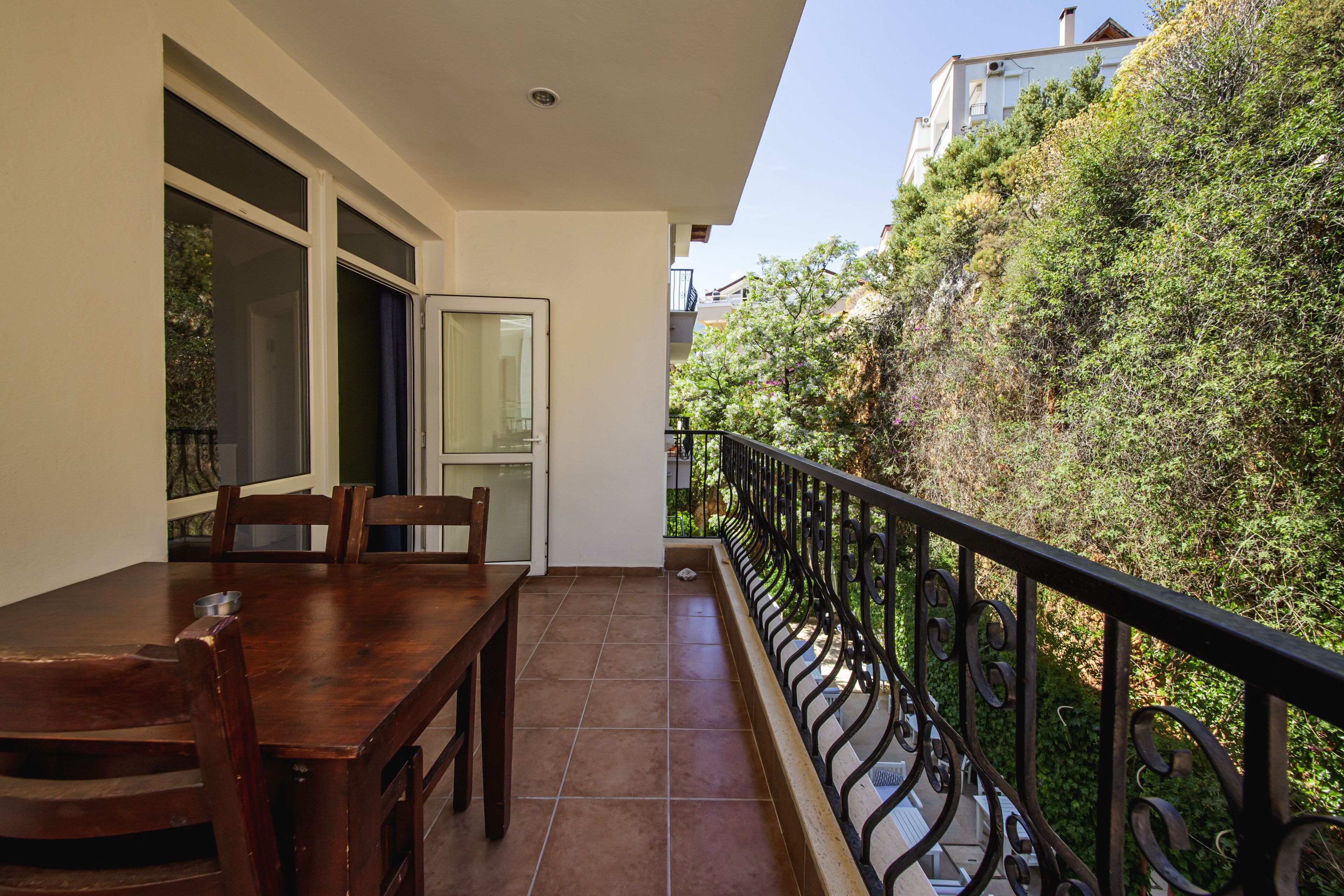 A balcony with a dining table and chairs, a view of a garden, and a white building in the background.