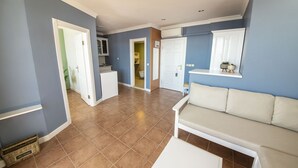Amplia sala de estar con un sofá color beige, una mesa de centro blanca, y una cocina visible a través de una puerta abierta.