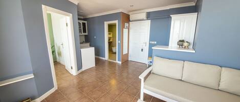 Amplia sala de estar con un sofá color beige, una mesa de centro blanca, y una cocina visible a través de una puerta abierta.