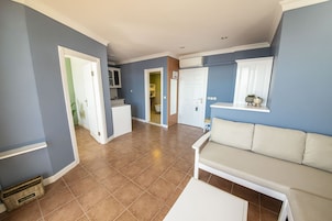 Amplia sala de estar con un sofá color beige, una mesa de centro blanca, y una cocina visible a través de una puerta abierta.