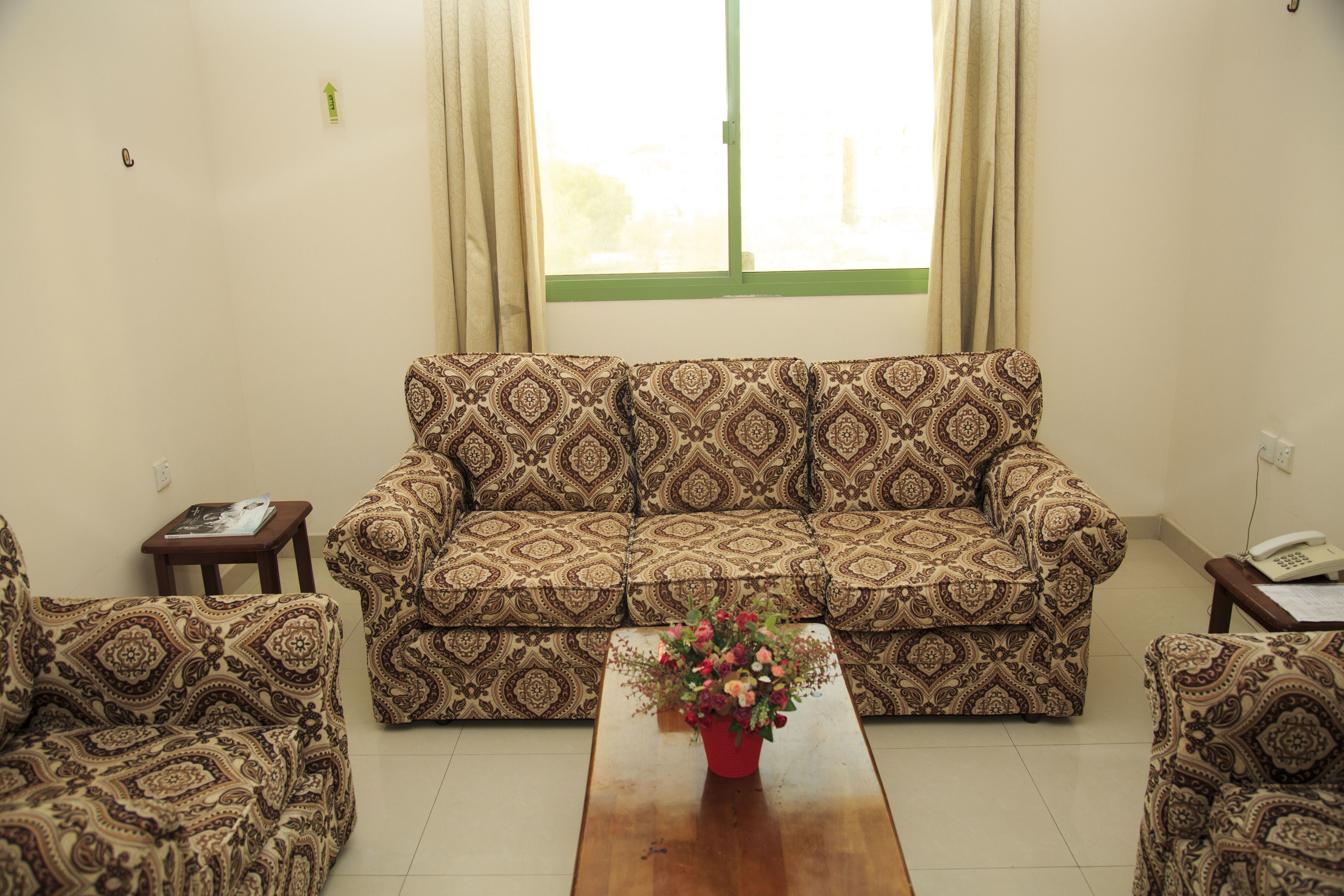 A living room with patterned sofas, a coffee table with flowers, a small side table, and a window with curtains.