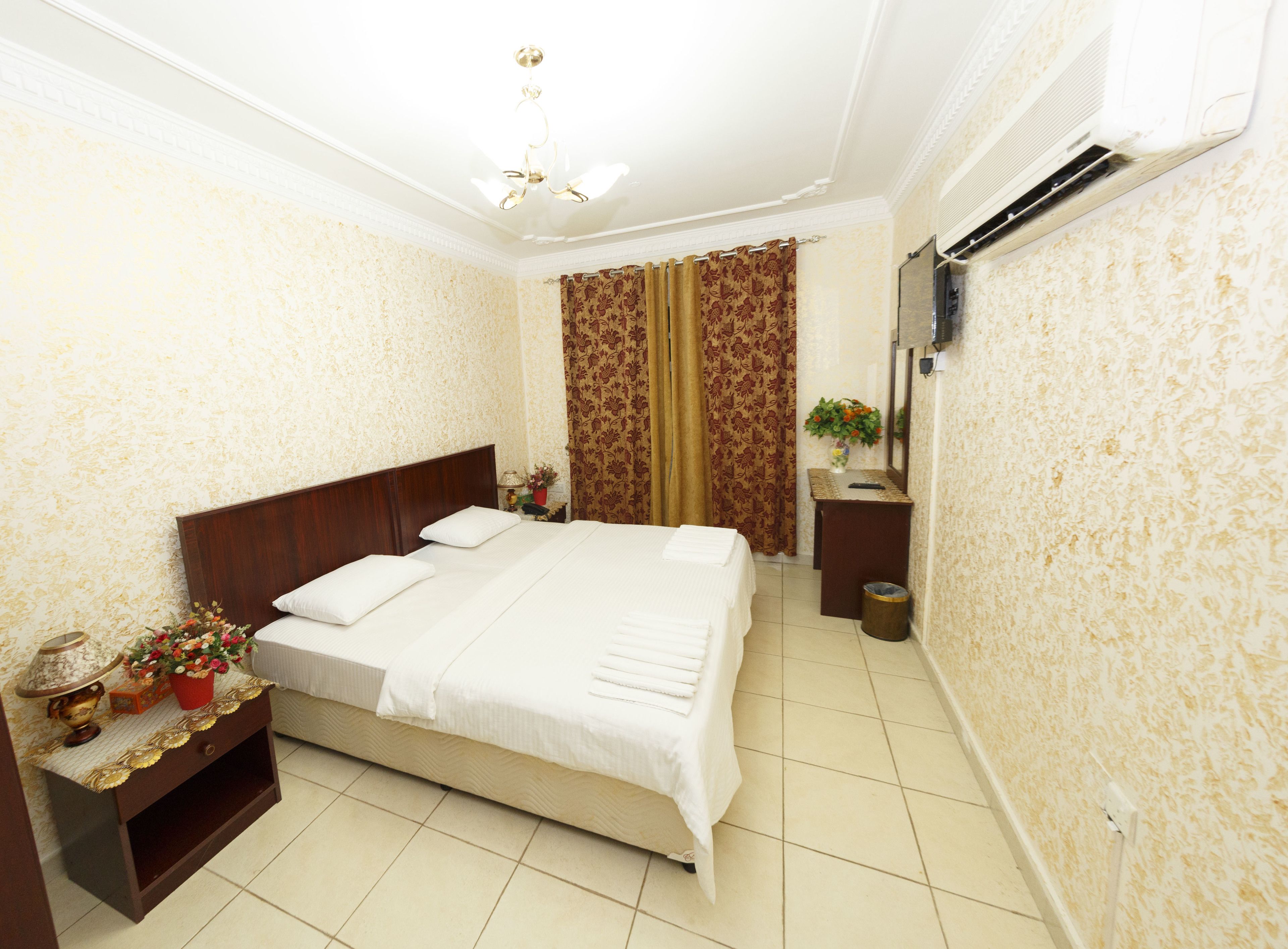 Habitación de hotel ordenada con una cama grande, una mesita de noche con lámpara, una pequeña mesa con un jarrón y un aire acondicionado de pared.