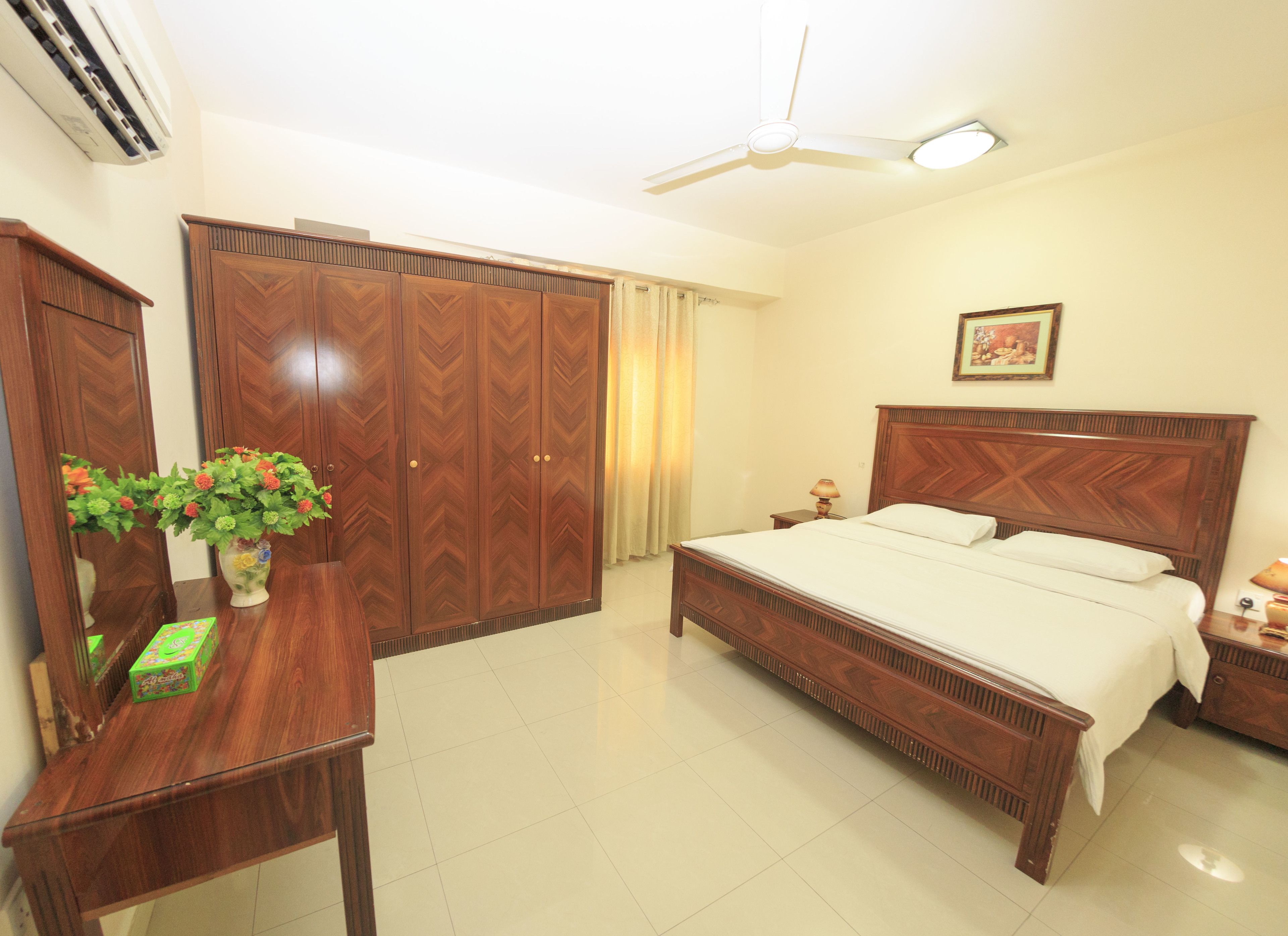 A bedroom with a bed, a wardrobe, a desk with a vase of flowers, and a ceiling fan.