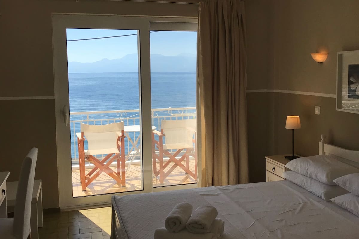 A hotel room with a bed, a nightstand, a lamp, a window with a view of the sea and mountains, and a chair by the window.