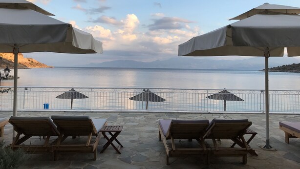 Eine Terrasse mit Holzliegen und Sonnenschirmen mit Blick auf ruhiges Wasser und entfernte Berge.