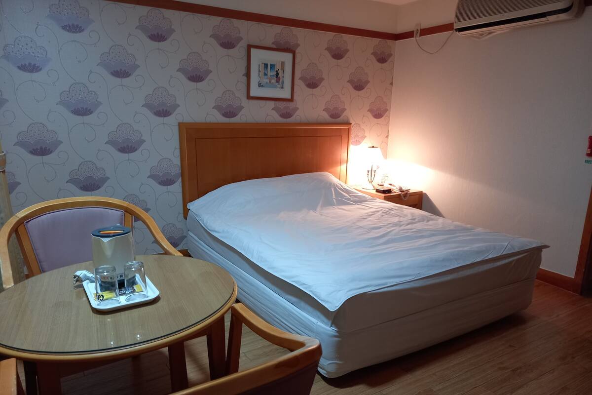 A hotel room with a bed, a bedside table with a lamp, a chair, and a table with a water bottle and glasses.