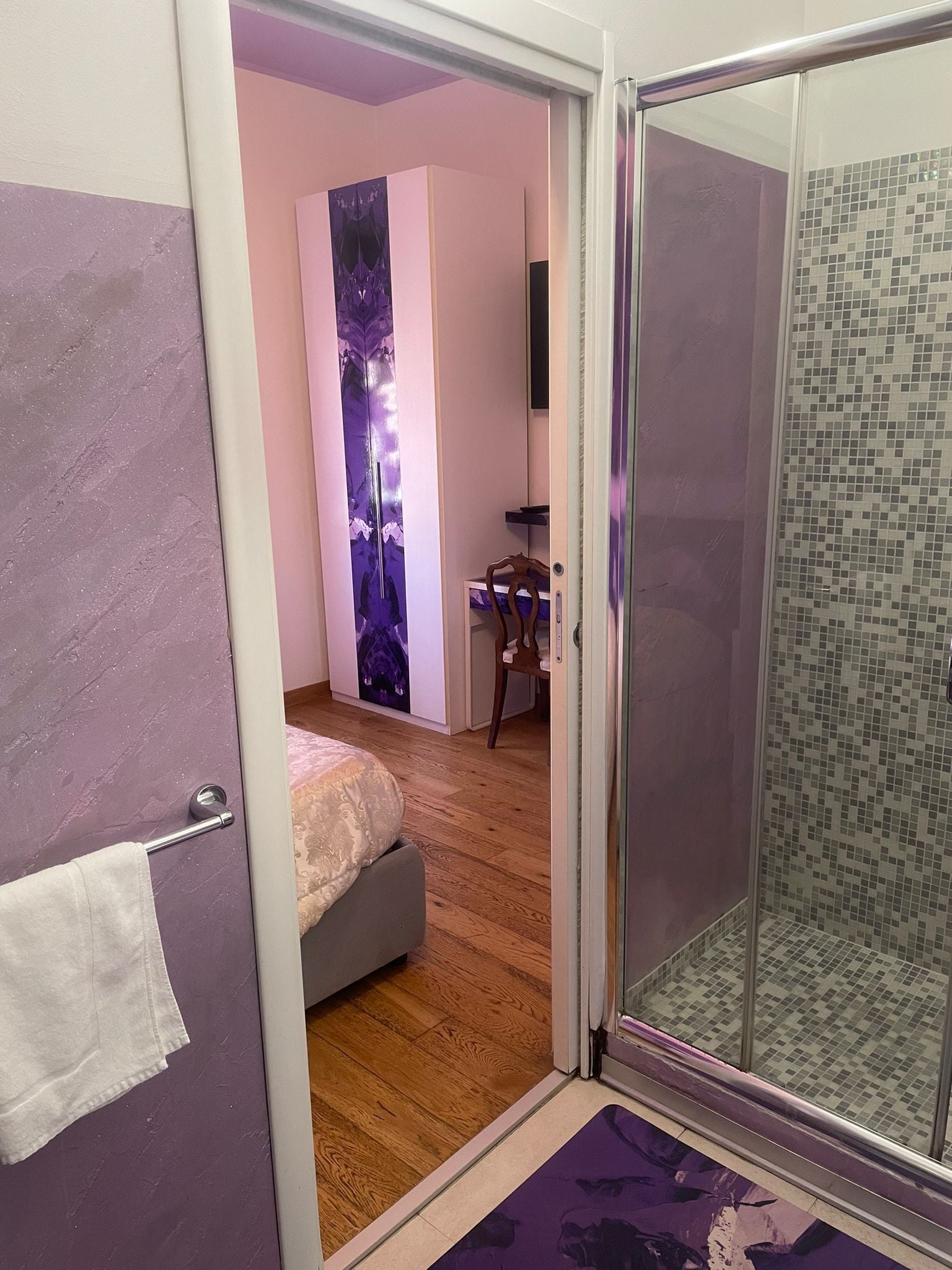 Una stanza con un letto, una scrivania, una sedia, box doccia in vetro e una parete viola decorata con motivi floreali.