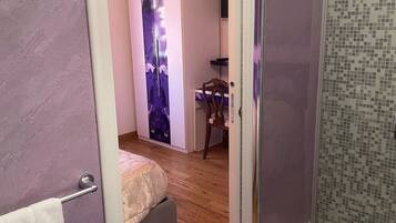 Una stanza con un letto, una scrivania, una sedia, box doccia in vetro e una parete viola decorata con motivi floreali.
