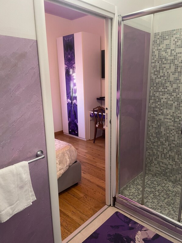 A room with a bed, a desk, a chair, a glass shower enclosure, and a purple wall with a floral design.