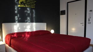 A bedroom with a red bedspread, white headboard, bedside lamps, and a wall with a poster. - Affittacamere Pavia (Pavia)