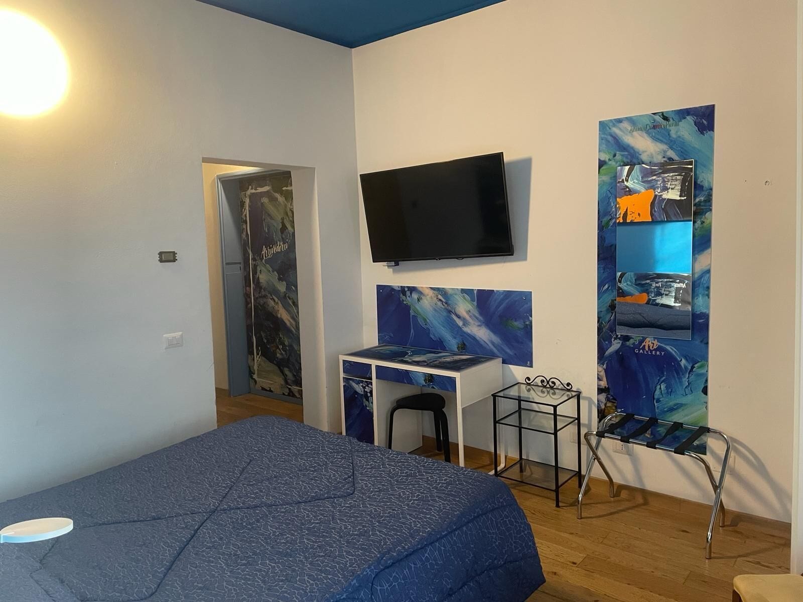A bedroom with a bed, a desk, a chair, a TV, and artwork on the walls.
