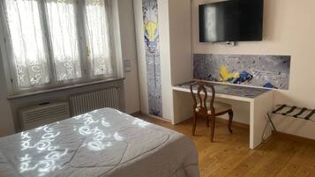 Ein Schlafzimmer mit einem Bett, einem Schreibtisch, einem Sessel, einem Heizkörper, einem Fernseher und einem Fenster mit Vorhängen.