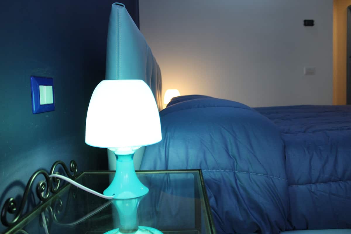 A room with a bed, a bedside table with a lamp, and a wall switch.