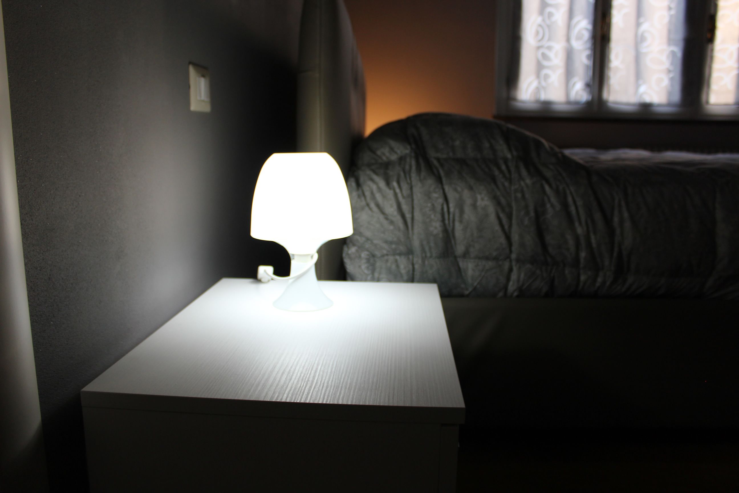 A bedside table with a lamp, a bed with a dark blanket, and a window with curtains.