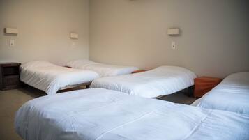 A room with four beds arranged in a row, each with white bedding. There are two wall-mounted light fixtures on either side of the beds.