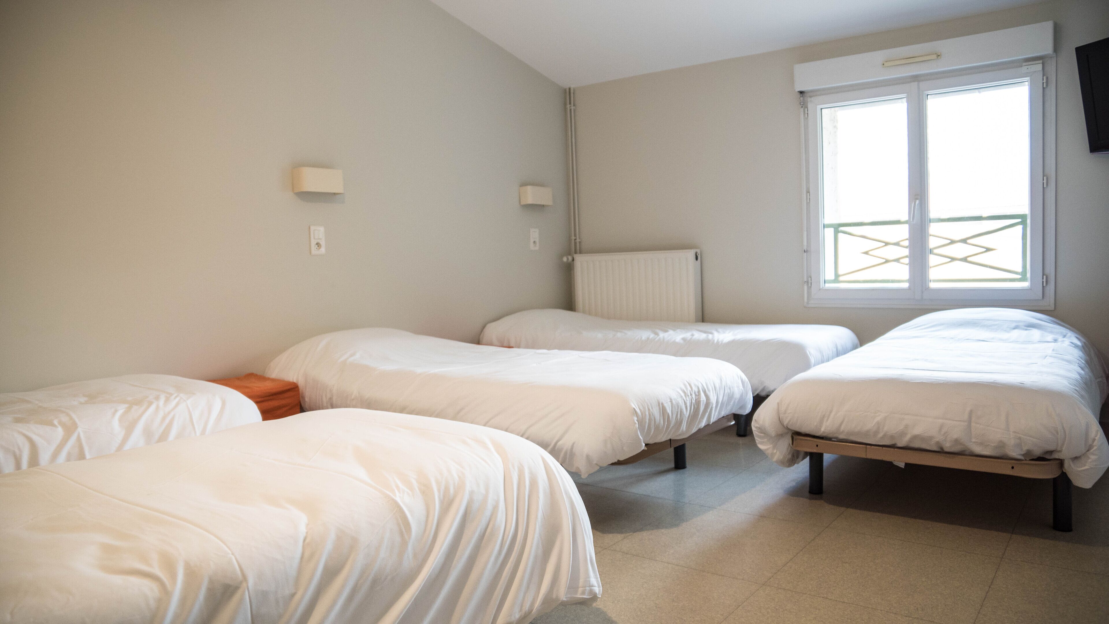 Een kamer met vier op een rij staande bedden, een radiator, een raam met uitzicht en twee aan de muur bevestigde lampen.