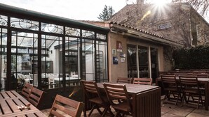 Un café al aire libre con mesas y sillas de madera, un cerramiento de cristal con barras metálicas y una montaña al fondo.