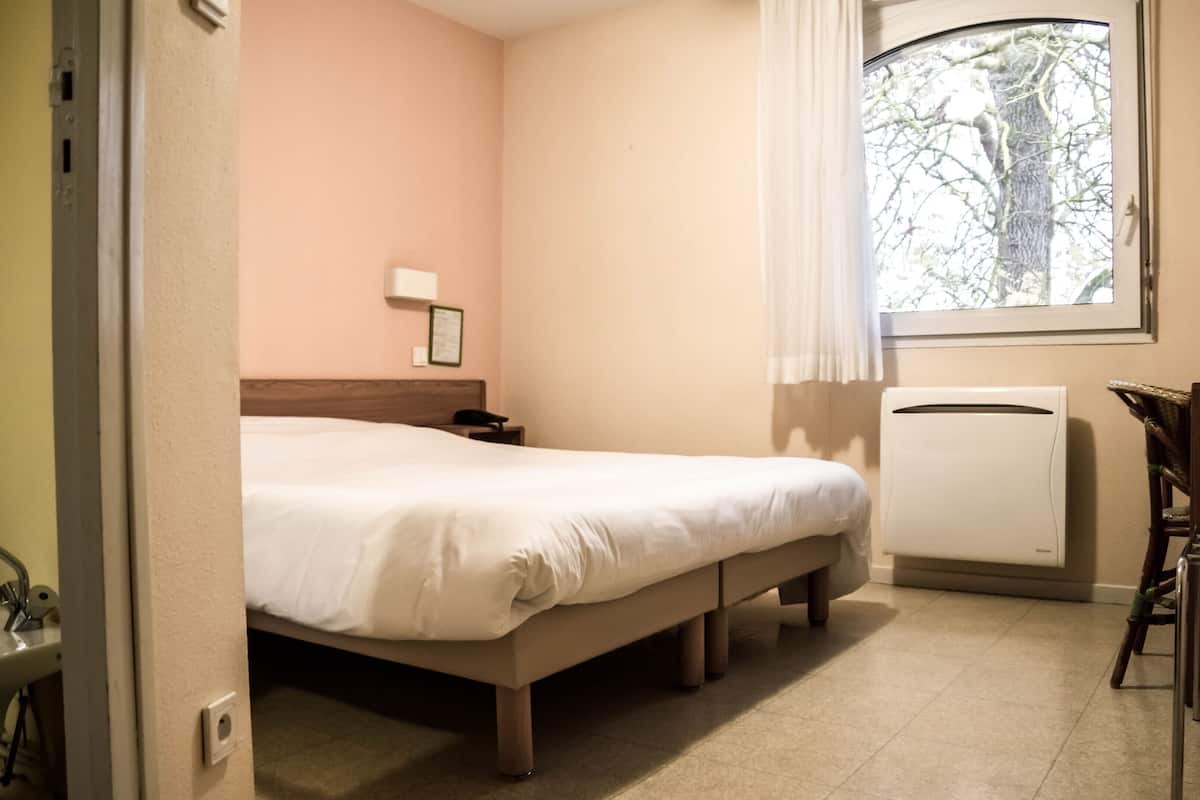Hotellihuone, jossa on sänky, yöpöytä, lamppu, patteri ja ikkuna, josta on näkymä puita kohti.