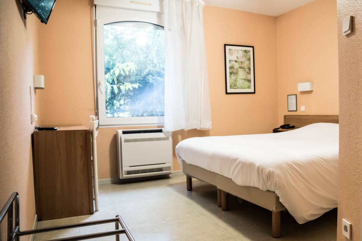 Hotellihuone, jossa on sänky, työpöytä, ikkuna verhoilla, ilmastointilaite ja seinällä oleva kehystetty kuva.