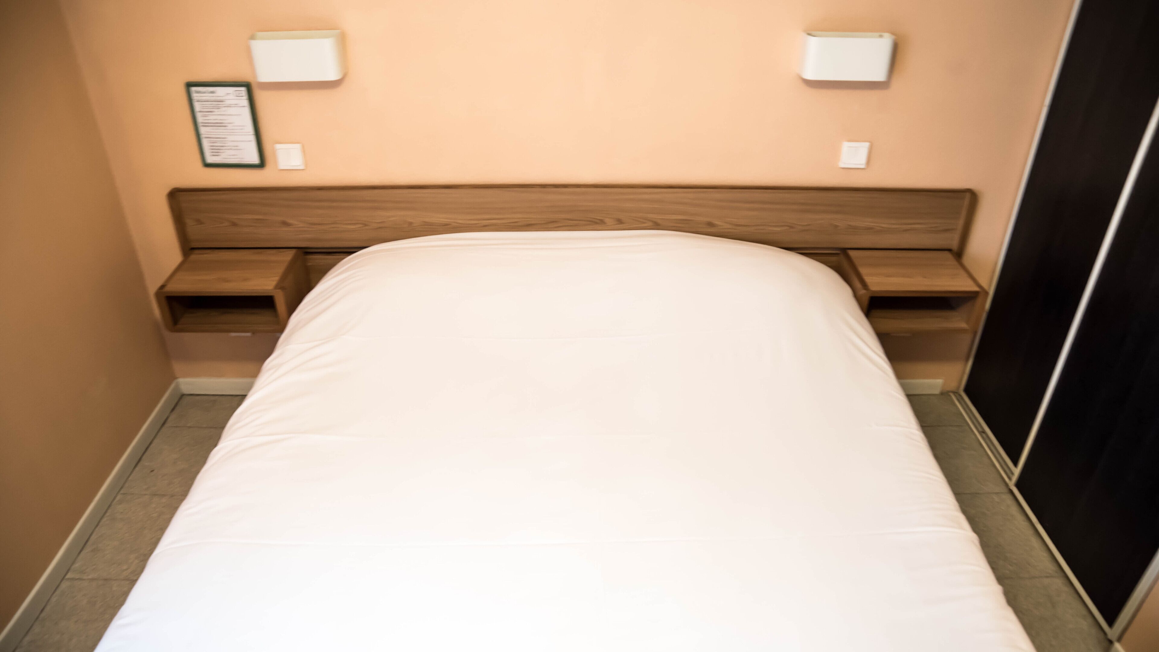 A hotel room with a single bed, wooden headboard, bedside tables, and wall-mounted light fixtures.