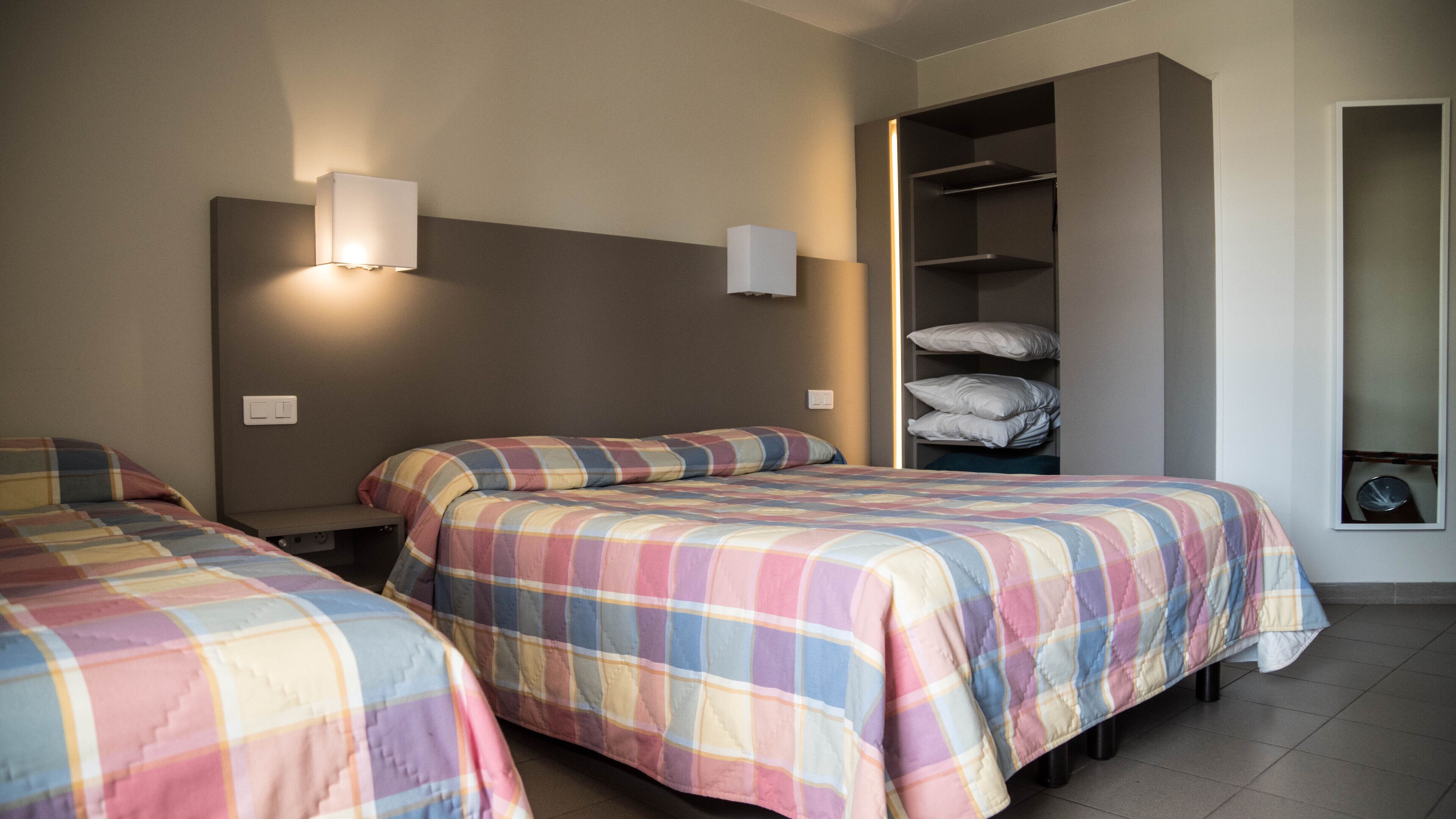 Habitación de hotel con dos camas, cada una con cubrecama a cuadros, cabecera, mesita de noche y lámpara de pared.