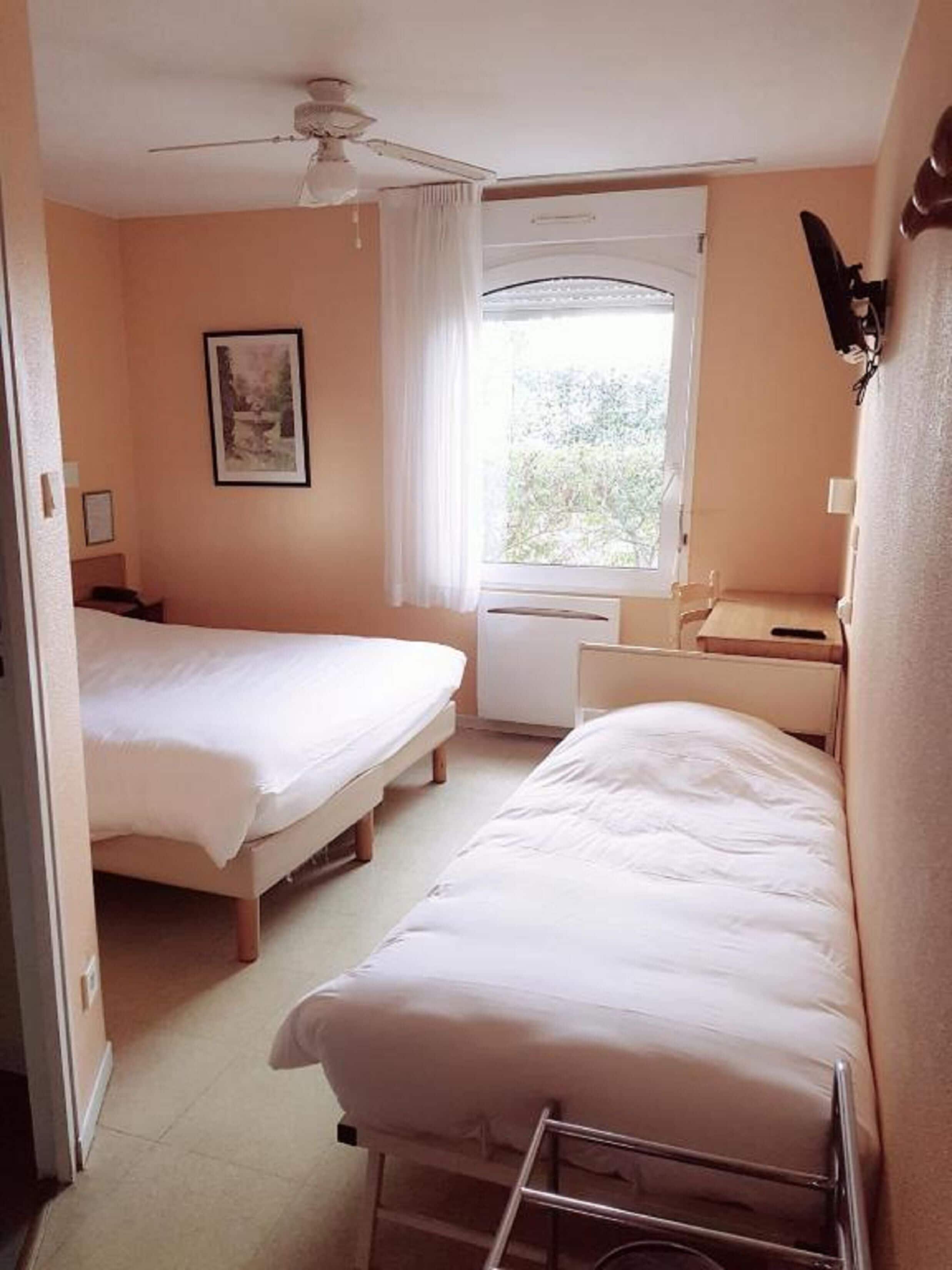Une chambre avec deux lits, un ventilateur de plafond, une fenêtre avec des rideaux et un petit bureau avec une chaise.