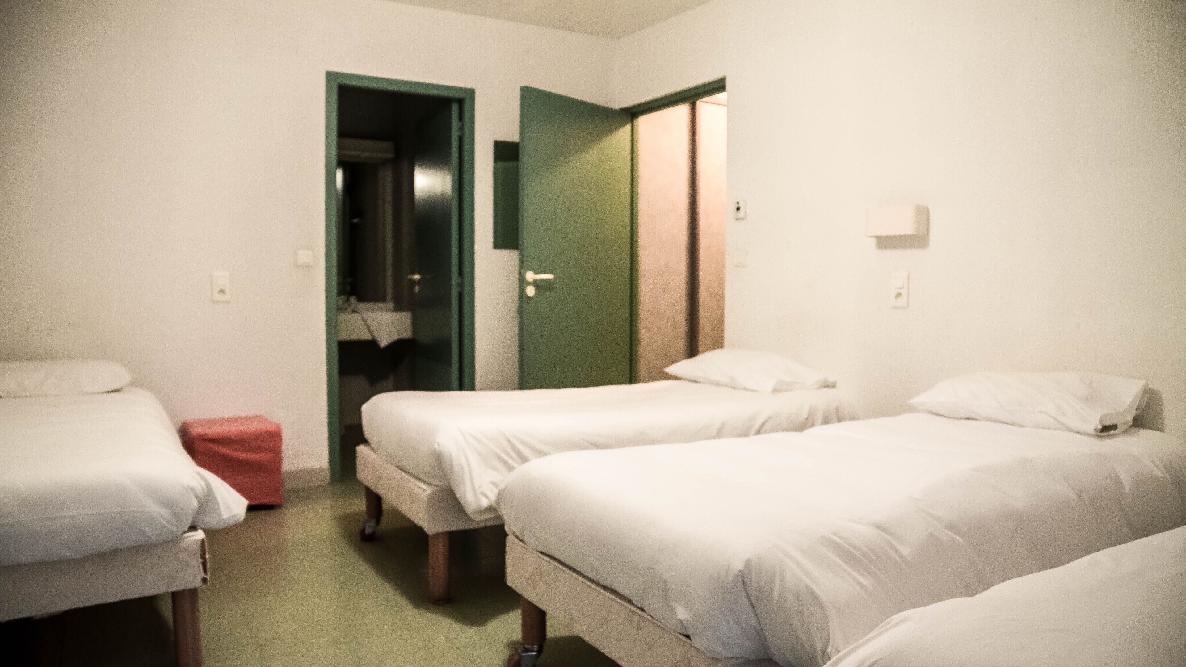 Quarto com três camas de solteiro, um banquinho vermelho pequeno e uma porta verde que dá para um banheiro.