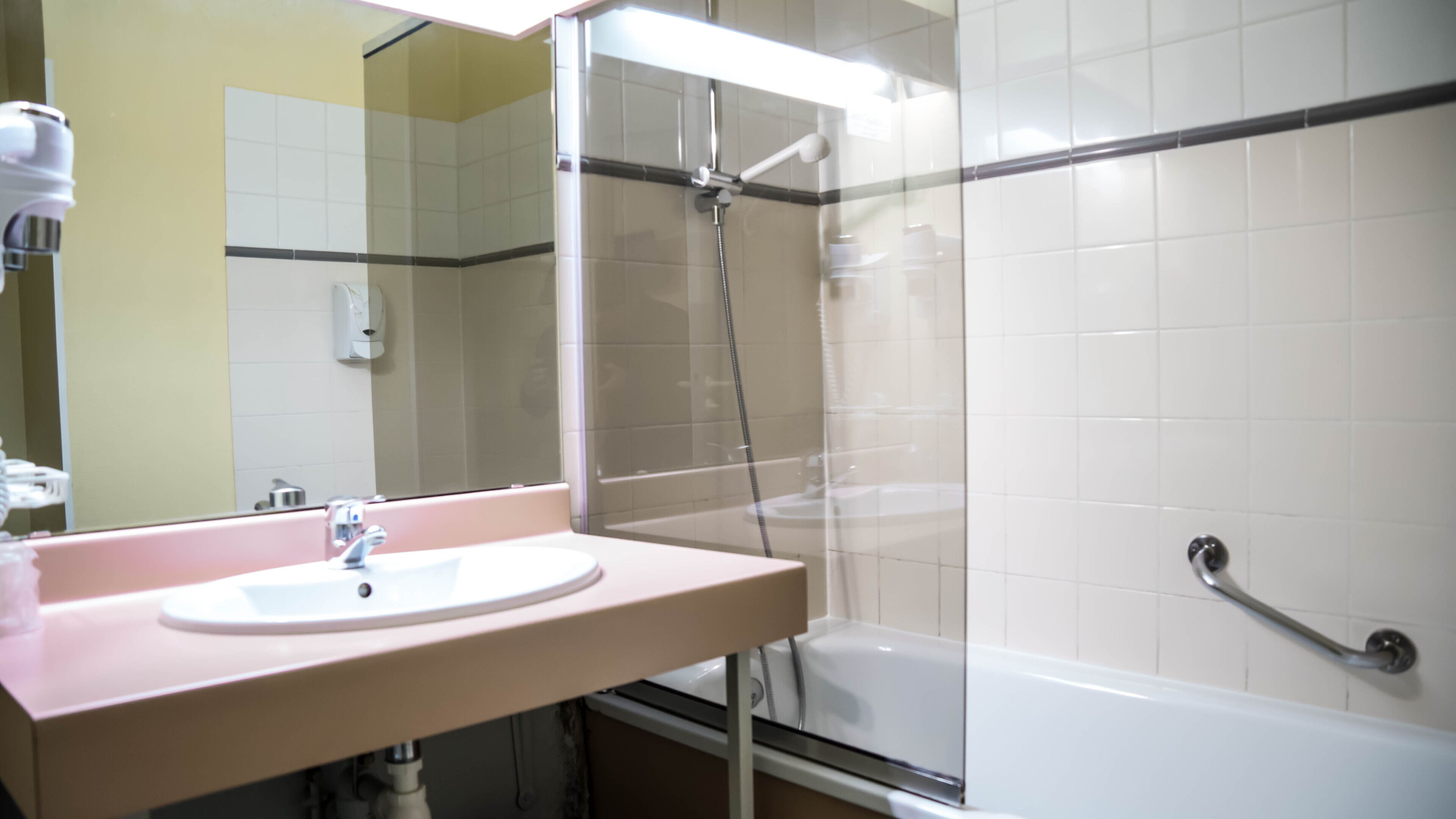 A bathroom with a sink, mirror, shower, and handrail.