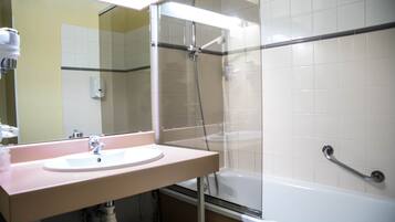 A bathroom with a sink, mirror, shower, and handrail.