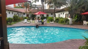 A swimming pool with lounge chairs and umbrellas, surrounded by greenery and buildings.