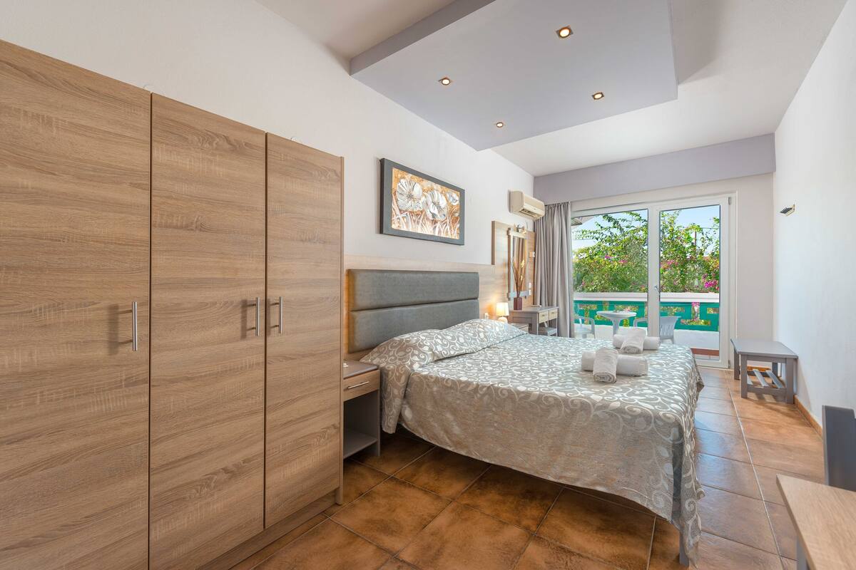 A bedroom with a large wardrobe, a bed with a patterned bedspread, a nightstand, and a view of a balcony with outdoor furniture.