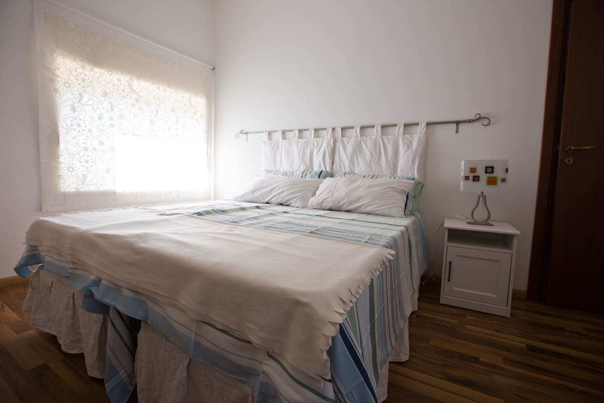 Un dormitorio con cama, mesita de noche, ventana con cortinas traslúcidas y una puerta.