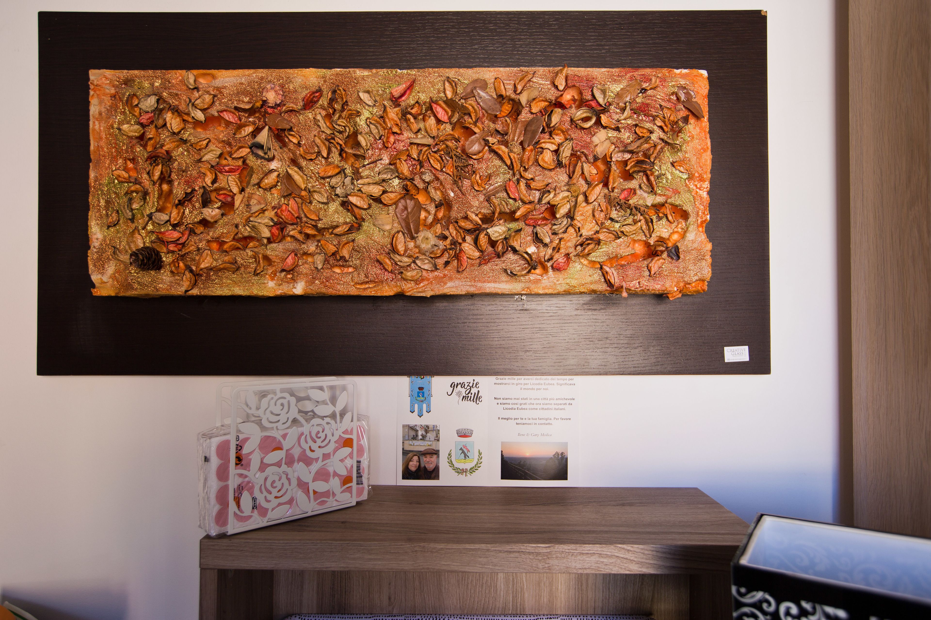 Una habitación con una obra de arte grande en la pared, una pequeña mesa con un adorno, y un certificado o premio enmarcado en la pared.