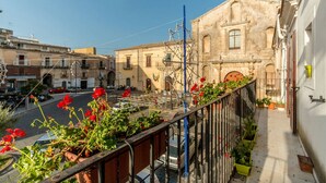 A narrow street with buildings on either side, a metal fence, and potted plants with red flowers. - Il Paesino (Licodia Eubea)