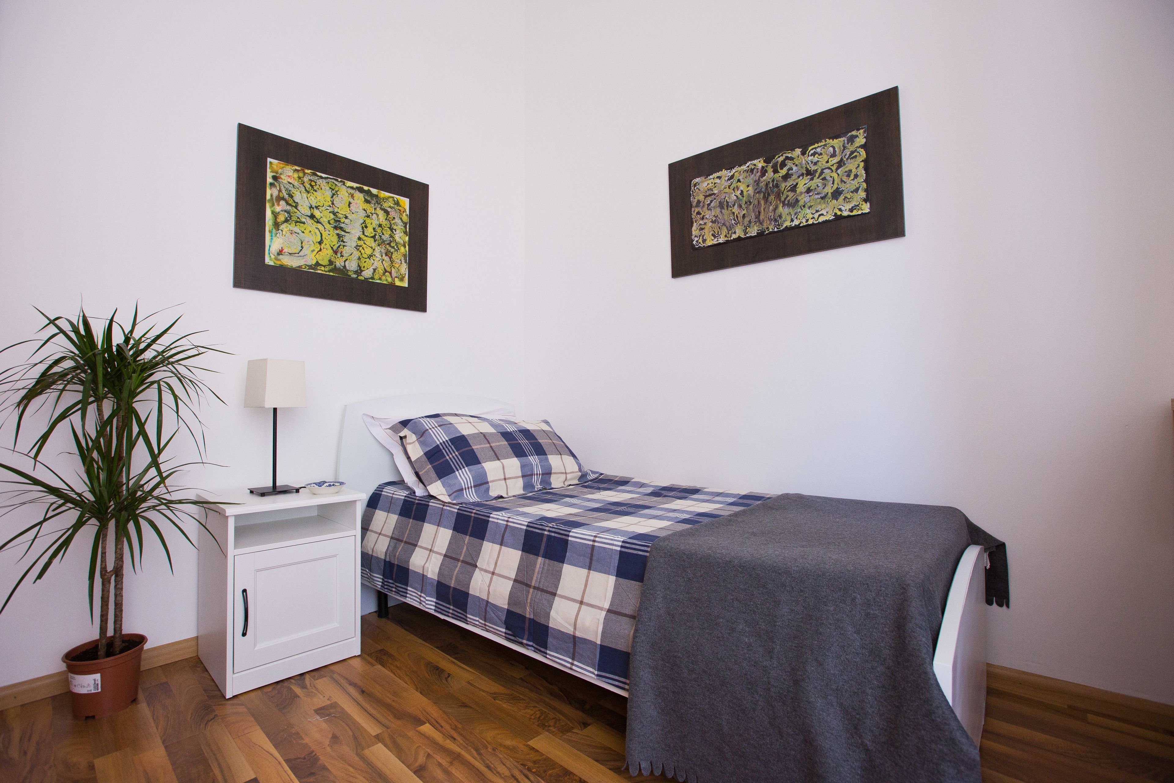 A bedroom with a bed, a nightstand, a lamp, a potted plant, and two abstract paintings on the wall.