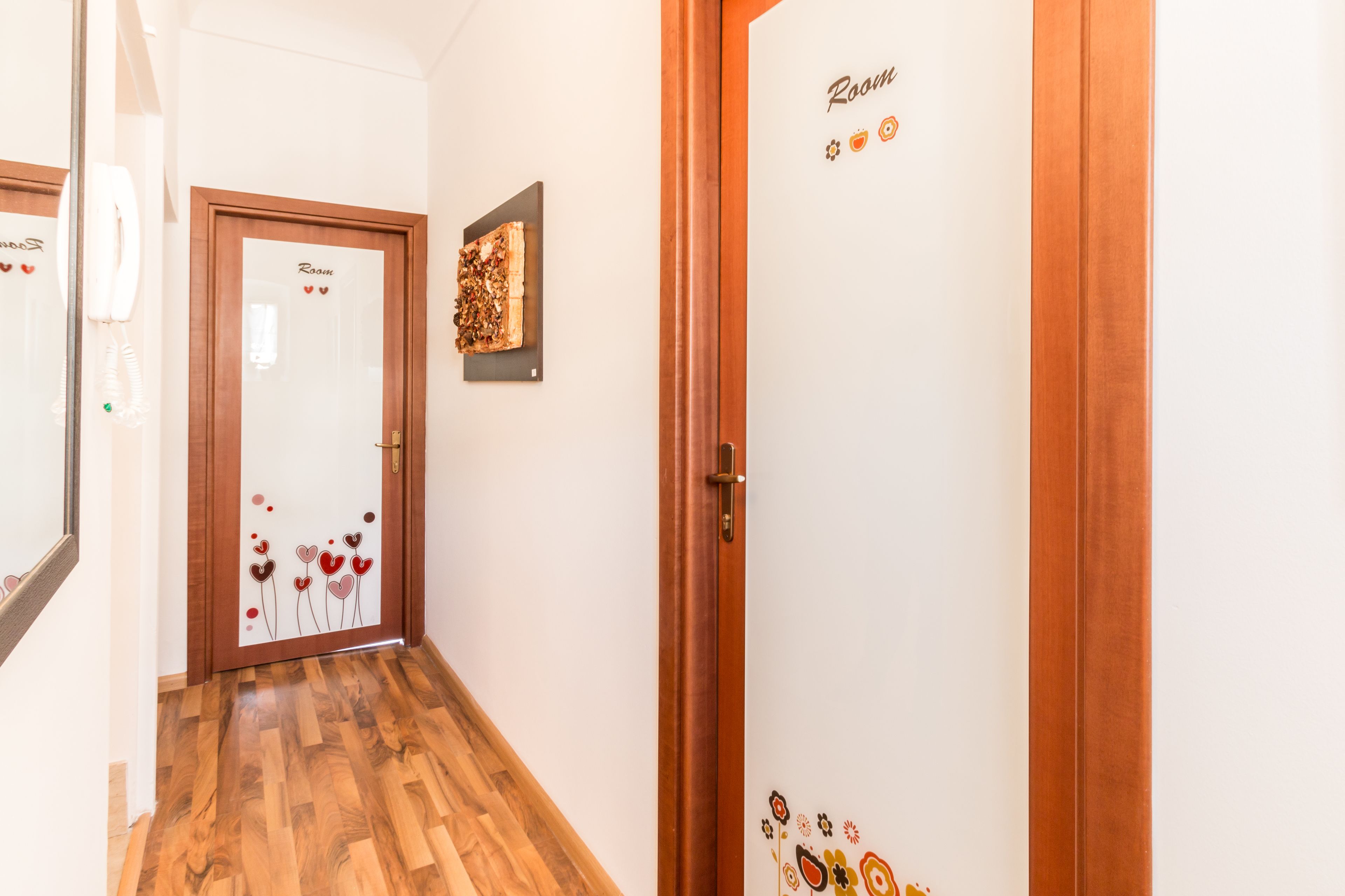 A hotel hallway with wooden flooring, a door labeled "Room," and a framed picture on the wall.