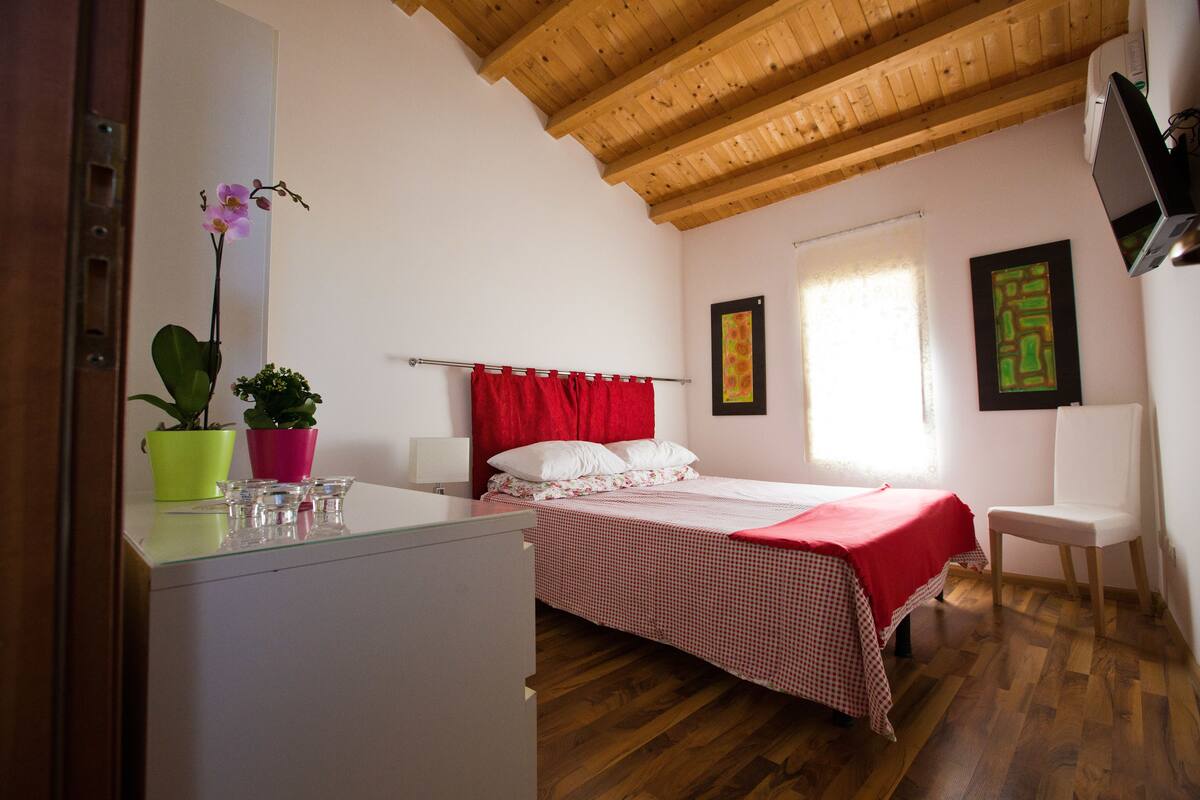 Un dormitorio con techo de madera, cama con cabecera roja, escritorio con planta en maceta y ventana con cortinas.