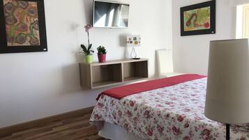 A hotel room with a wooden floor, a bed with a floral bedspread, a TV mounted on the wall, and two potted plants on a shelf.