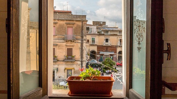 A window with a potted plant, a view of a street with buildings, and a door with a handle. - Il Paesino (Licodia Eubea)