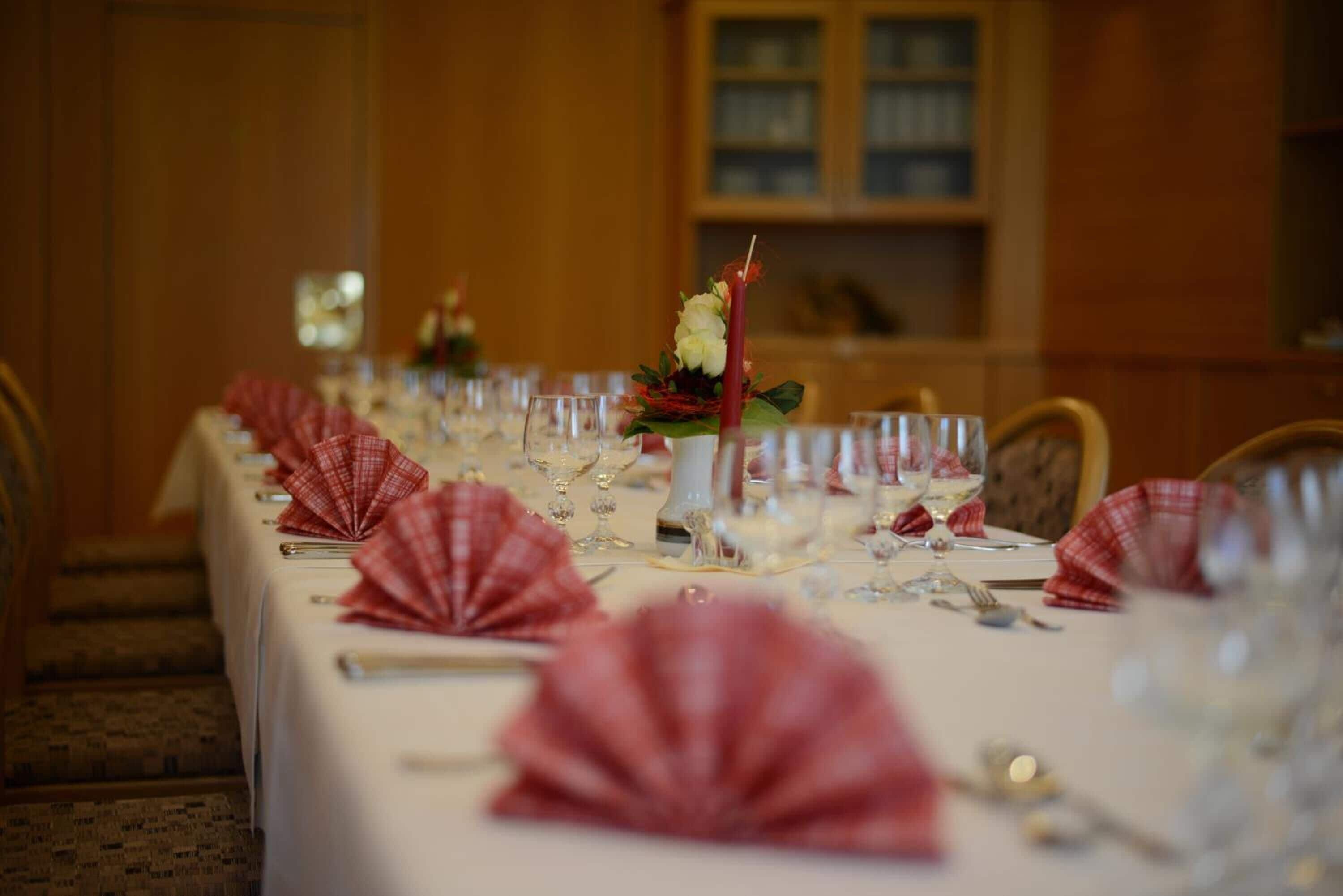 Une longue table à manger, dressée avec des serviettes rouges, des verres à vin et un centre de table composé de fleurs et d’une bougie.
