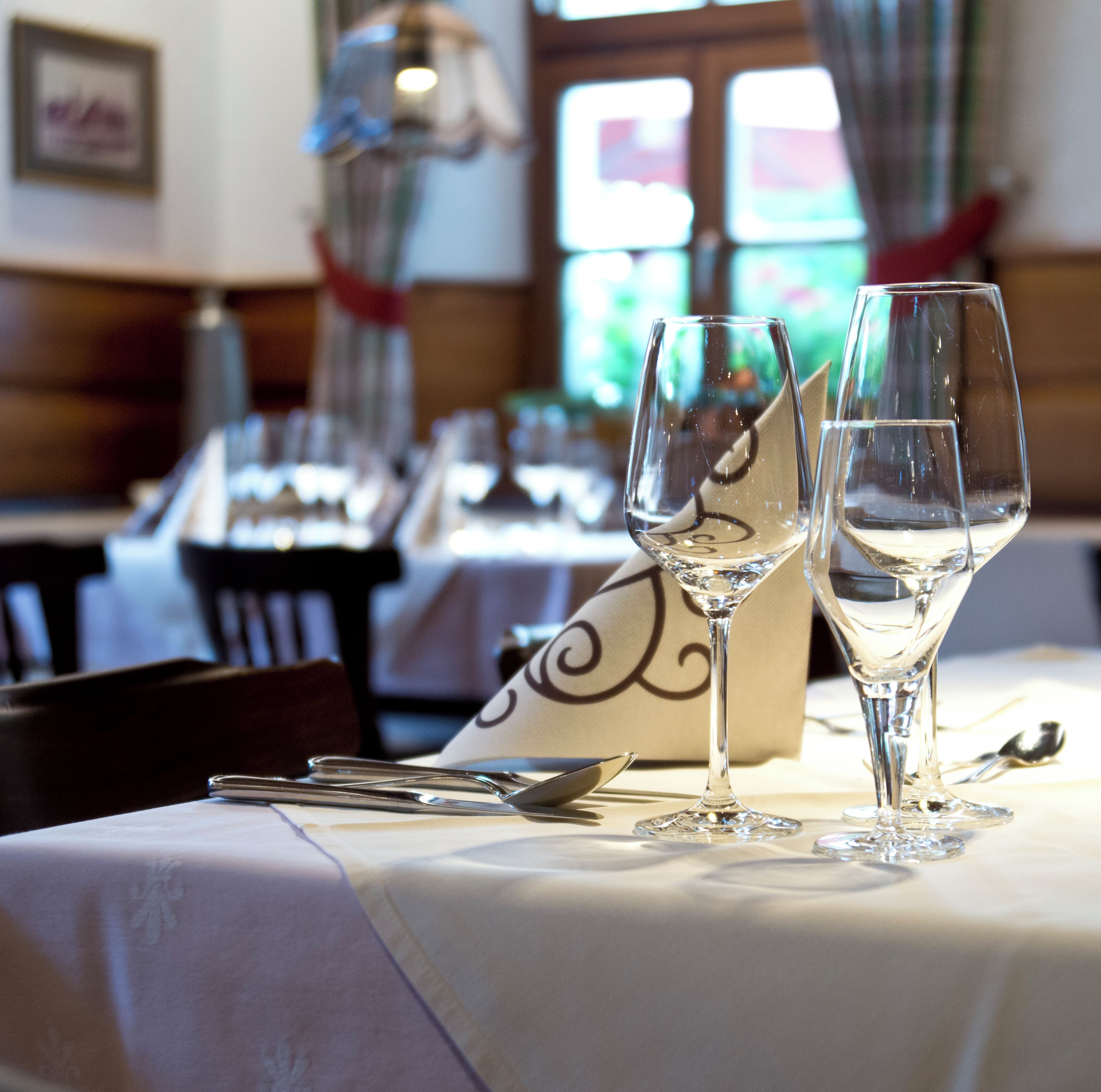 Une table à manger avec deux verres à vin et des couverts, un coussin décoratif et une fenêtre avec des rideaux, le tout dans un décor de restaurant.