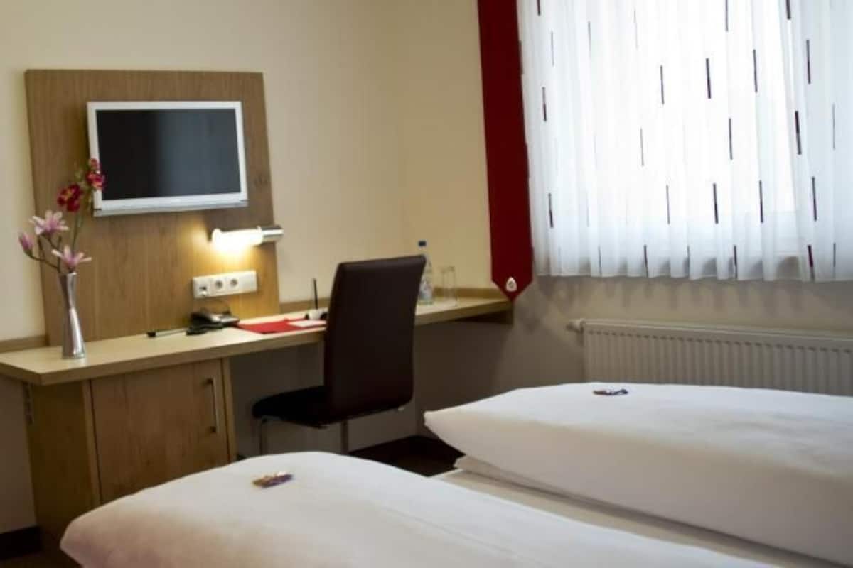 Une chambre d’hôtel avec deux lits, un bureau avec une chaise, une télévision et un vase avec des fleurs.