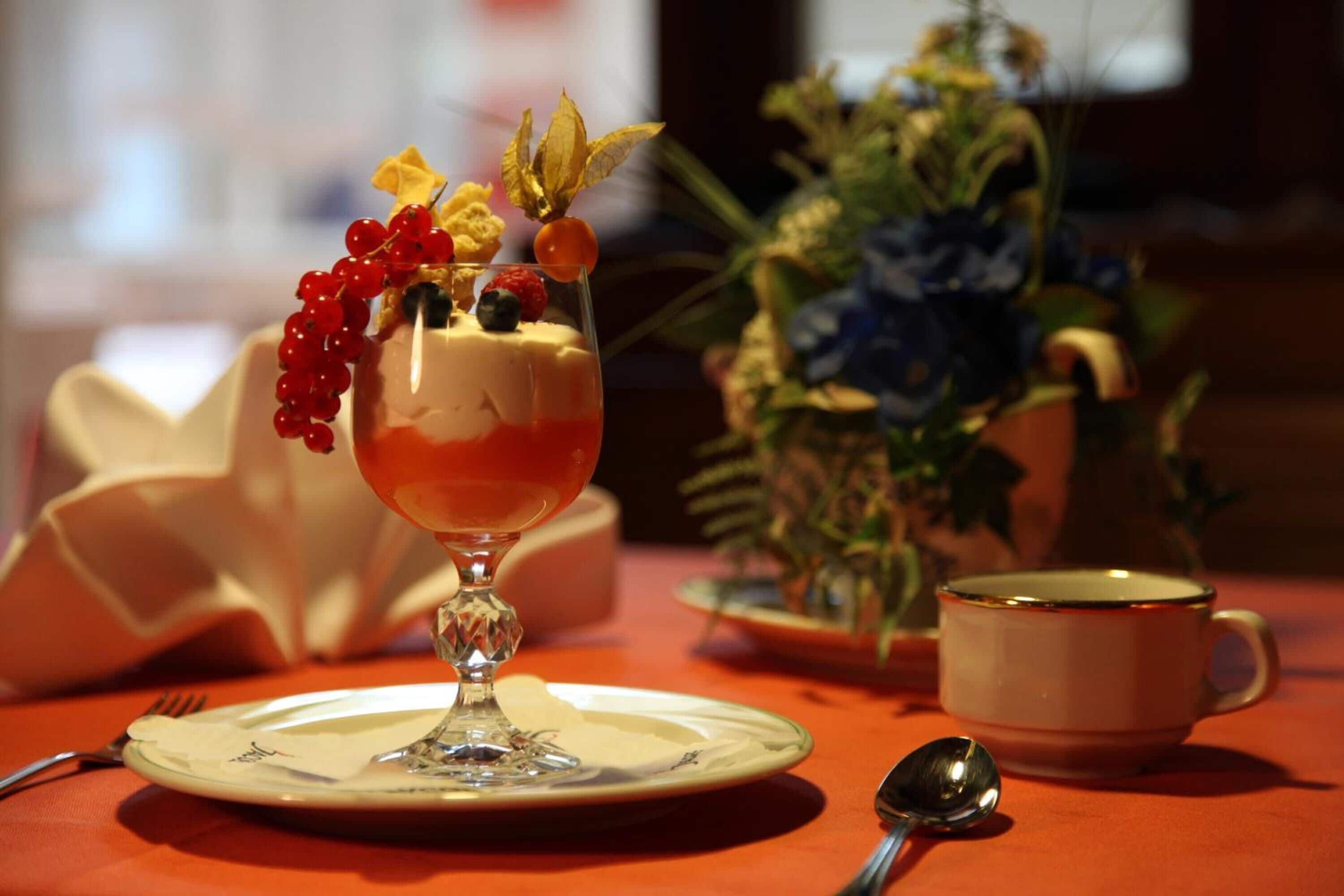 Dessert i et glas på en tallerken, en kop og en ske på et bord med en plante i baggrunden.