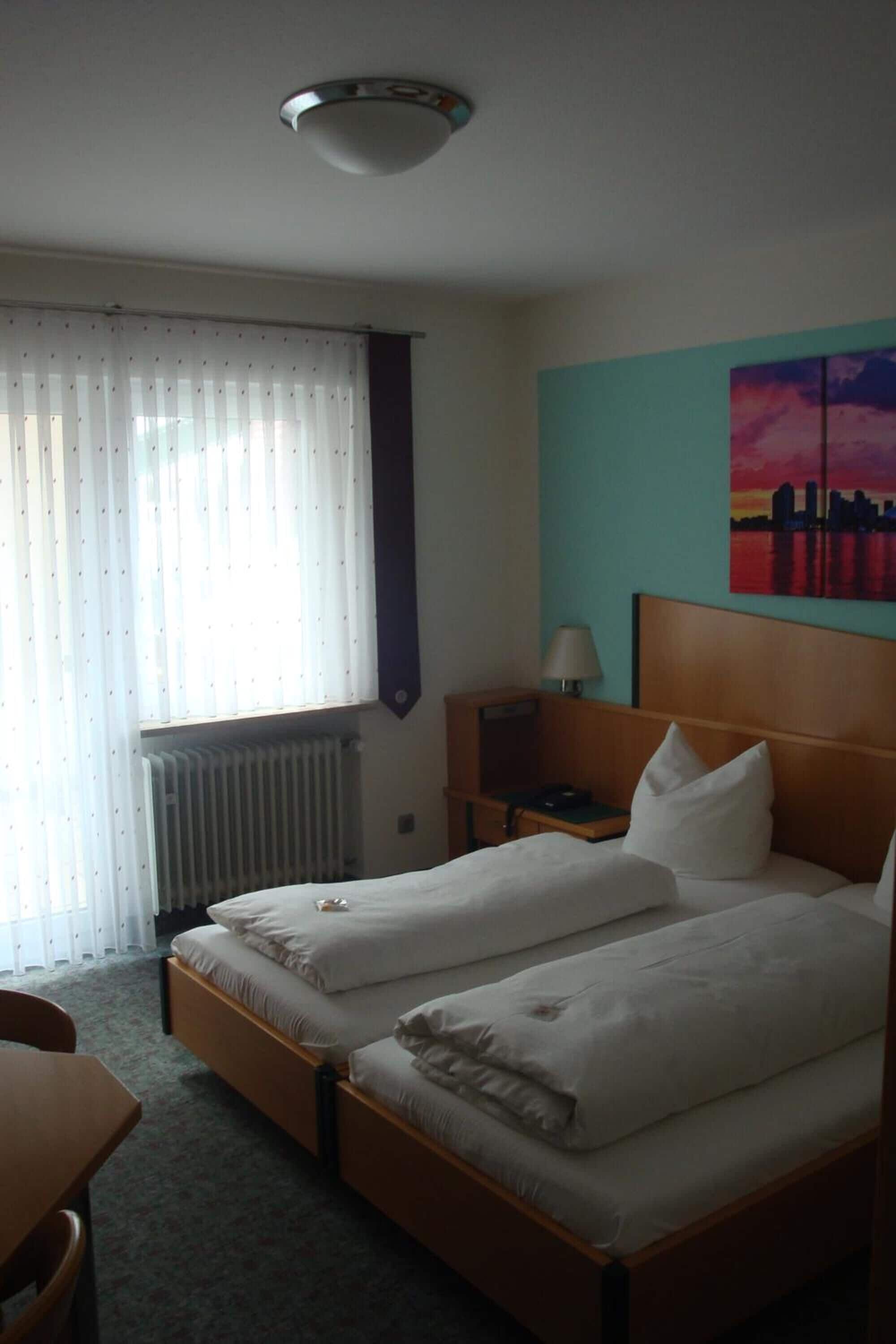 Een hotelkamer met twee eenpersoonsbedden, een bureau, een stoel, een radiator, een nachtkastje met een lamp en een stadsgezicht schilderij aan de muur.