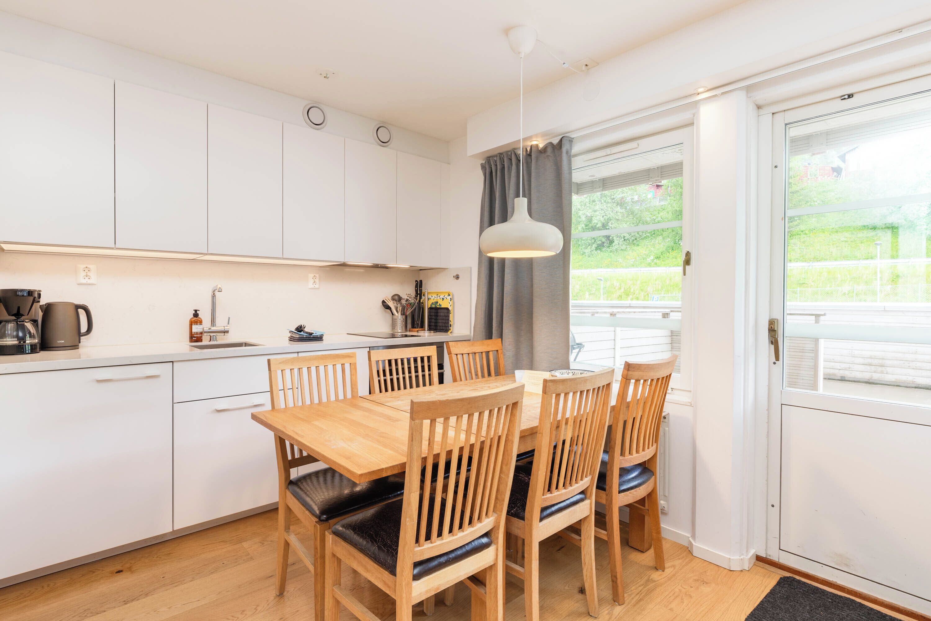 A modern kitchen with white cabinets, a wooden dining table with chairs, a coffee maker, and a window with a view of greenery.